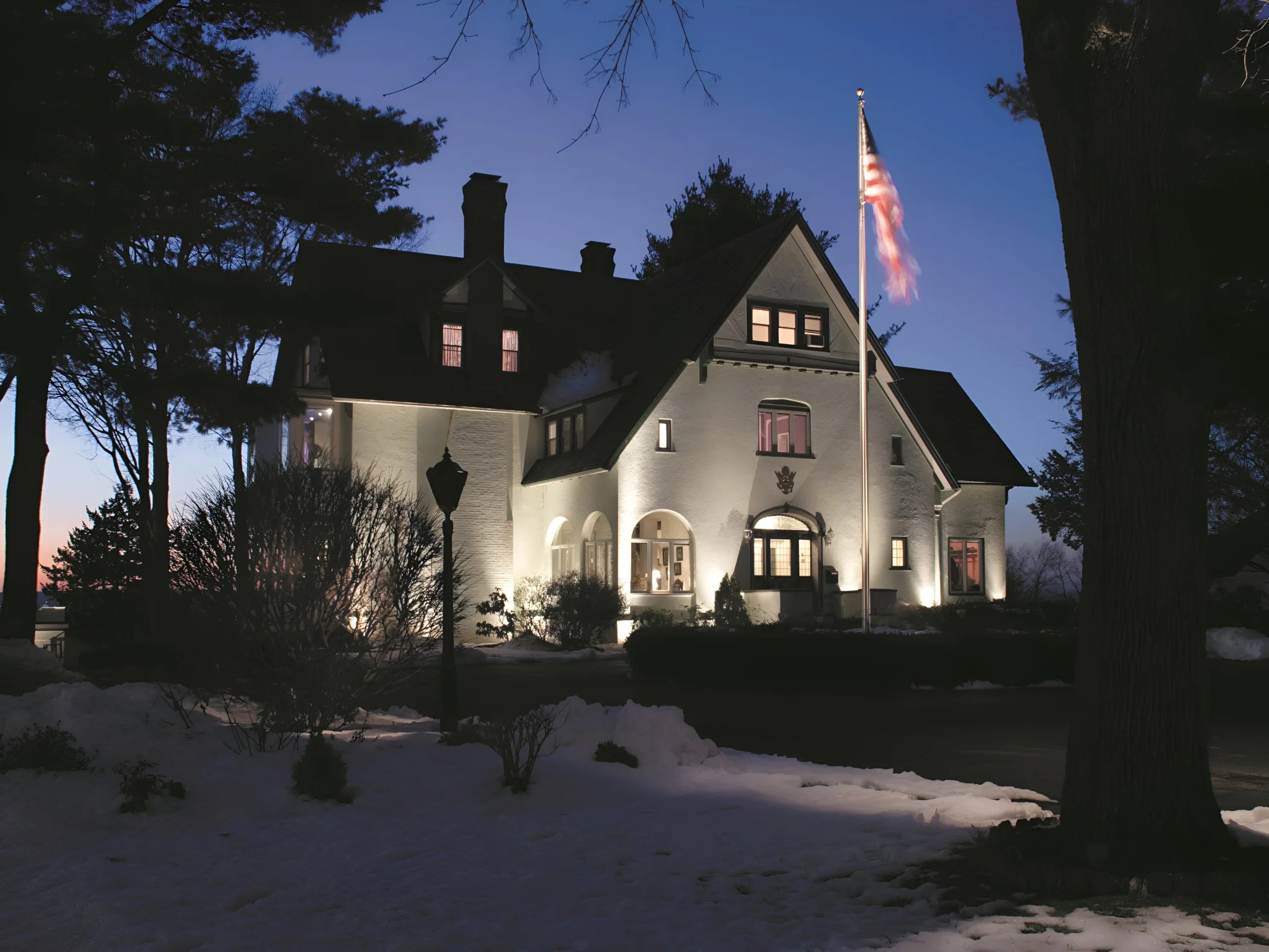 Uplights wash the pale walls of a large house and a flagpole beside a snowy lawn.