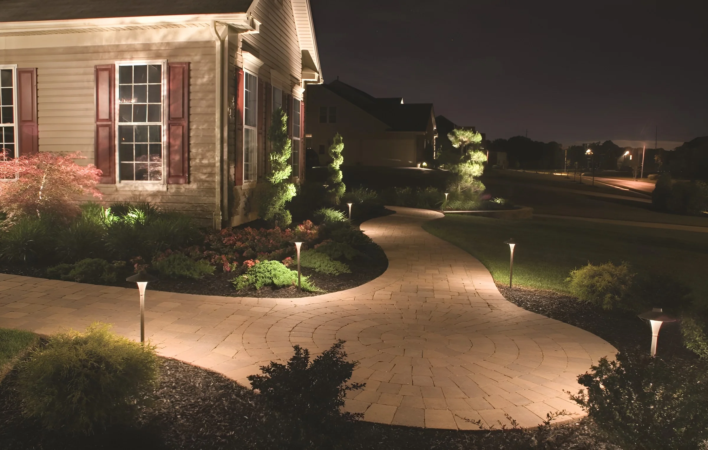 Path lights stagger along a wide curving front walk past lit planting beds and a brick house at night.