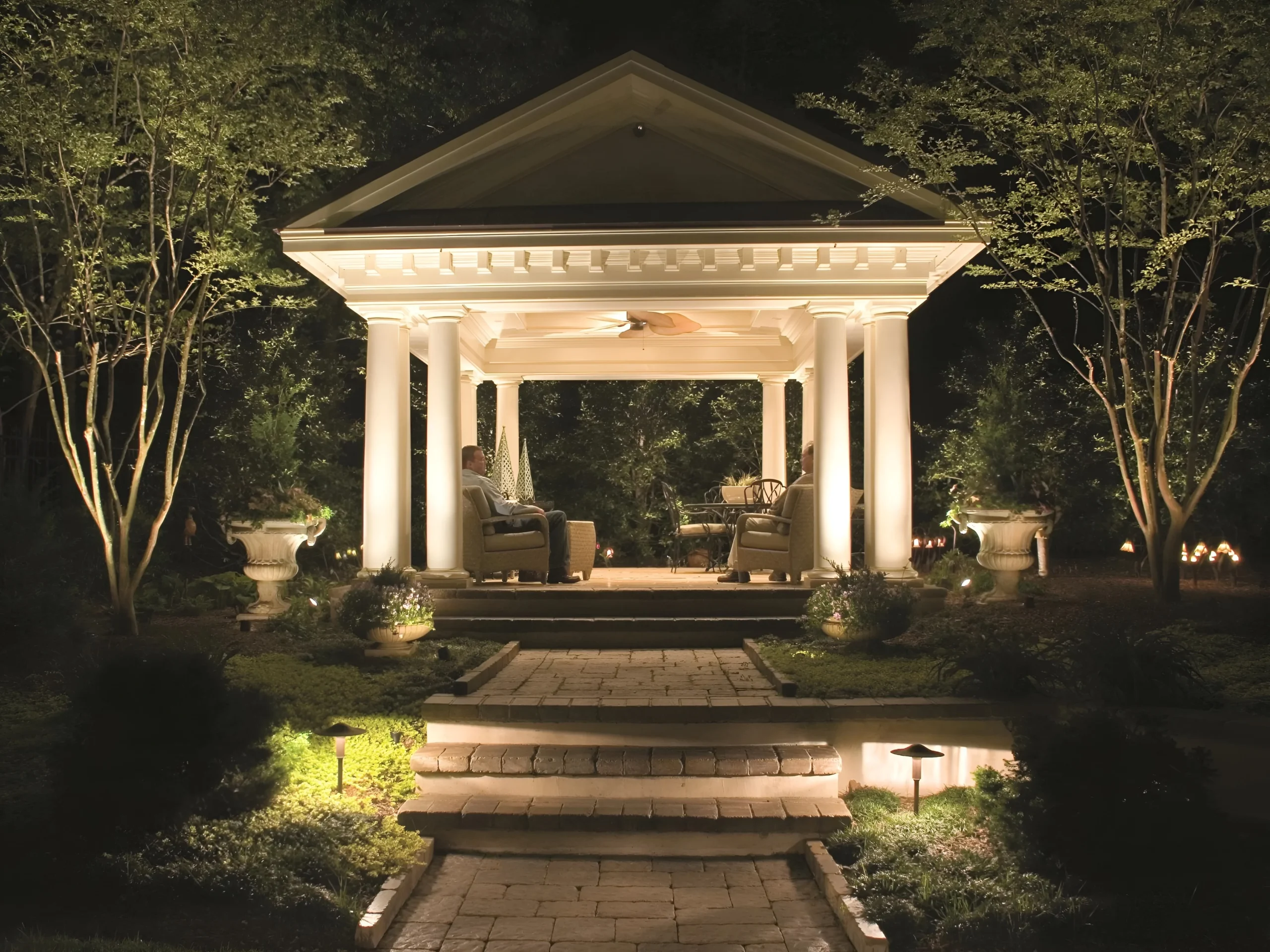 Uplit trees and path lights lead along a paver walk to a lit columned garden pavilion.